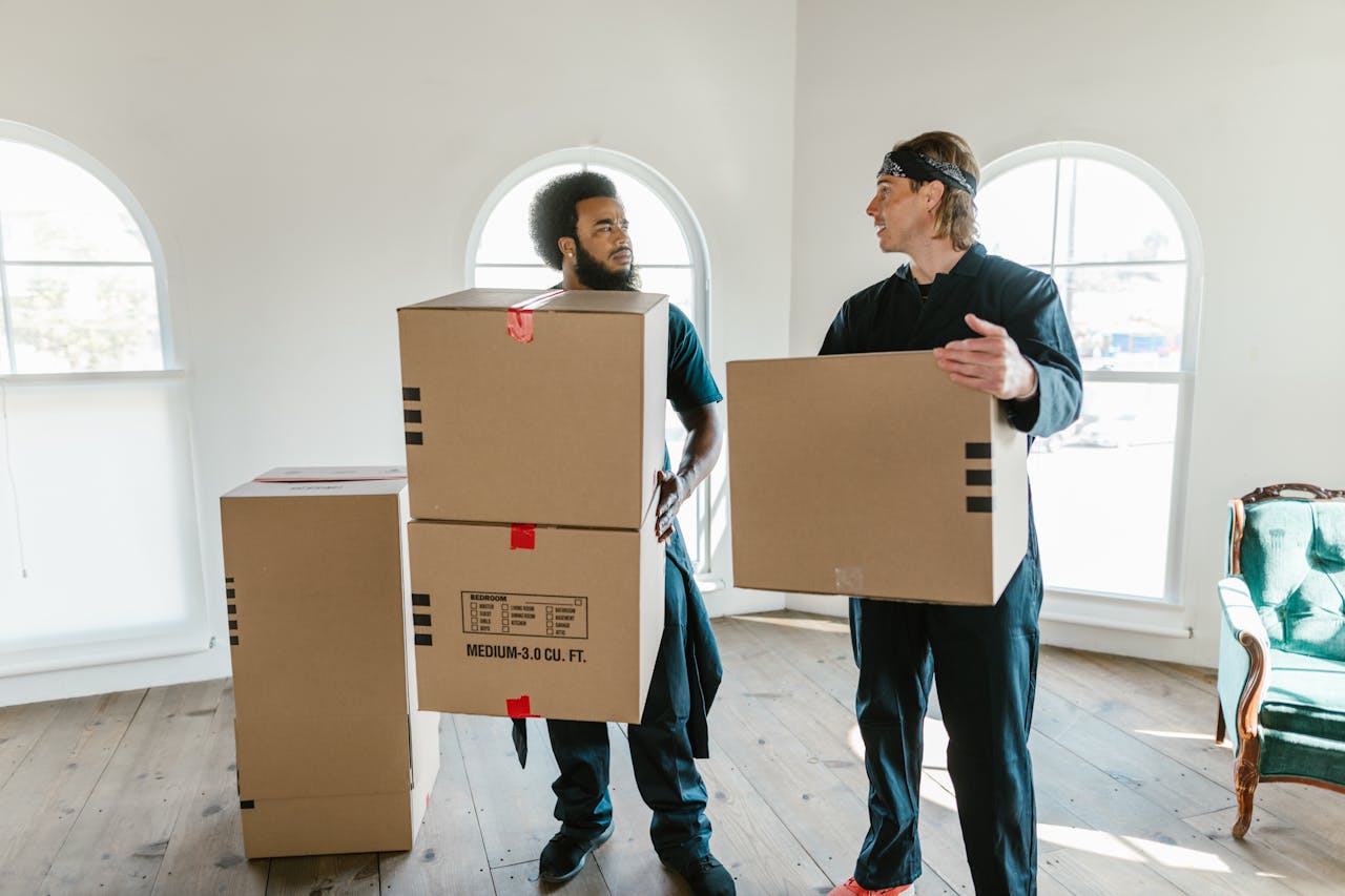 who-we-are Two professional movers carrying cardboard boxes inside a bright room with large windows.
